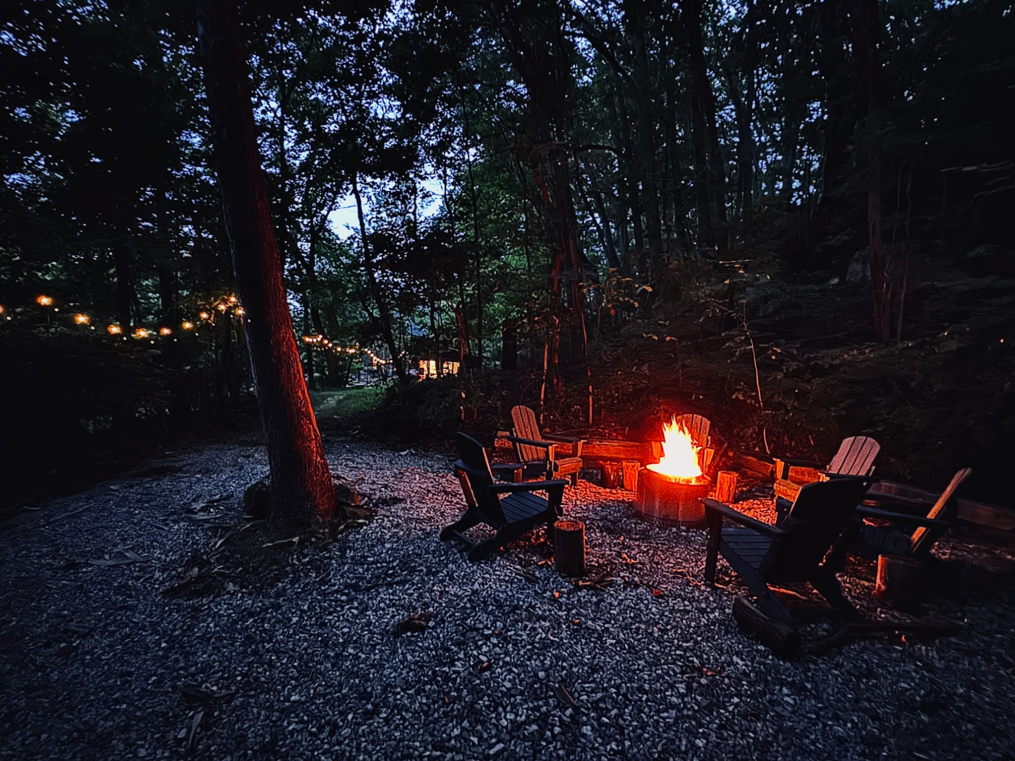 A fire burning in the fire pit at dusk, Adirondack chairs circled around it and string lights in the trees beyond