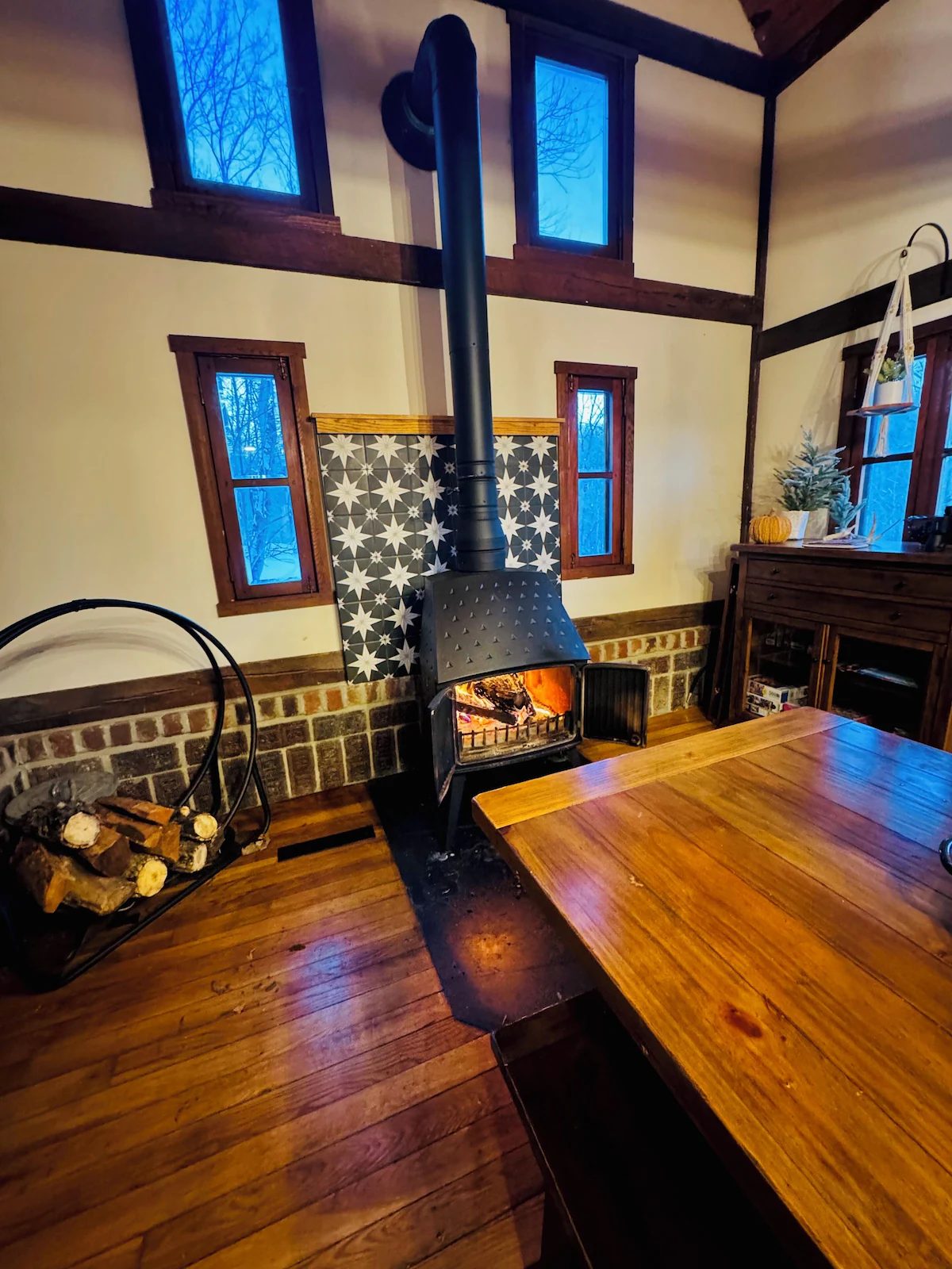 The wood stove lit at dusk, seen across the dining table with firewood stacked beside it